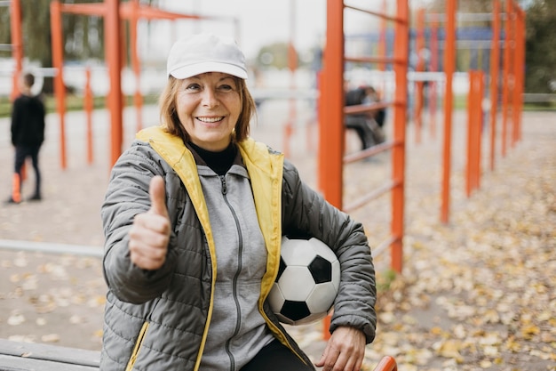 Mujer mayor sonriente sosteniendo el fútbol y dando pulgares mientras hace ejercicio