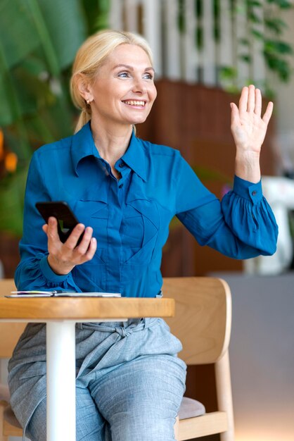 Mujer mayor sonriente sosteniendo smartphone y pidiendo algo mientras trabaja