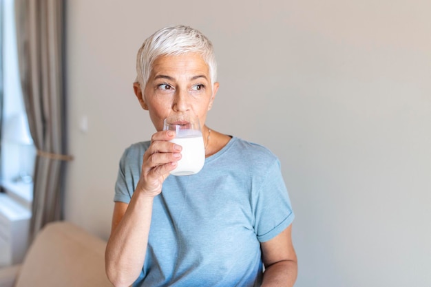 Mujer mayor sosteniendo un vaso de leche en casa Mujer de mediana edad bebiendo un vaso de leche fresca con una cara feliz de pie y sonriendo con una sonrisa segura mostrando los dientes