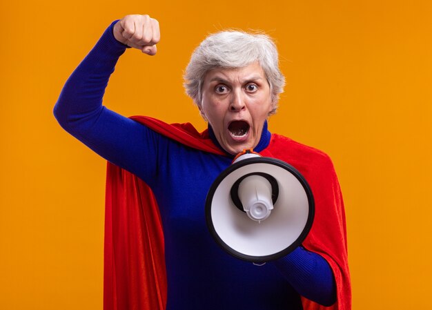 Mujer mayor superhéroe vistiendo capa roja gritando al megáfono con el puño cerrado de pie sobre fondo naranja