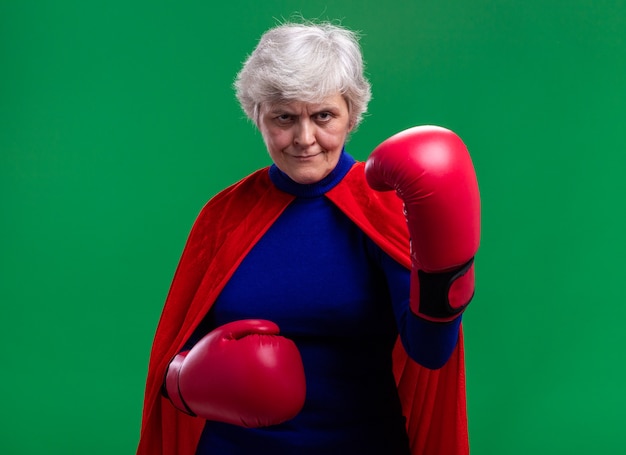Mujer mayor superhéroe vistiendo capa roja con guantes de boxeo mirando a la cámara con seria expresión de confianza lista para luchar
