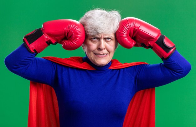 Mujer mayor superhéroe vistiendo capa roja con guantes de boxeo rodando los ojos hacia arriba está molesto e irritado de pie sobre fondo verde