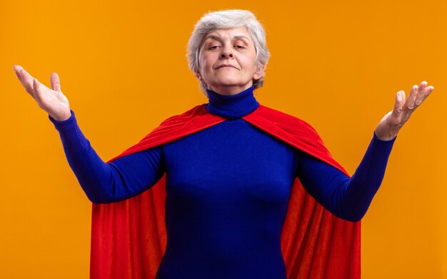 Mujer mayor superhéroe vistiendo capa roja mirando a la cámara con expresión de confianza con los brazos levantados sobre naranja