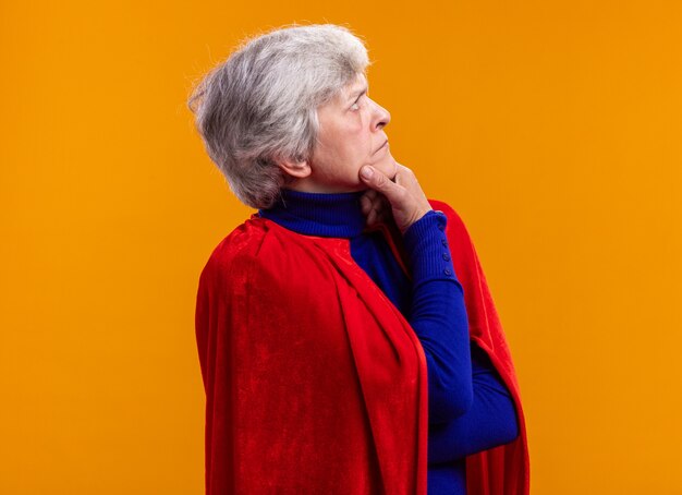 Mujer mayor superhéroe vistiendo capa roja mirando a un lado con rostro serio sobre naranja