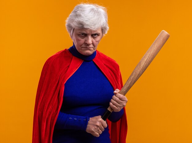 Mujer mayor superhéroe vistiendo capa roja sosteniendo un bate de béisbol mirando a la cámara con el ceño fruncido de pie sobre fondo naranja