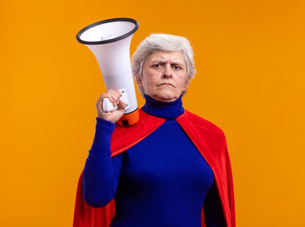 Mujer mayor superhéroe vistiendo capa roja sosteniendo megáfono mirando a la cámara con expresión de confianza de pie sobre fondo naranja