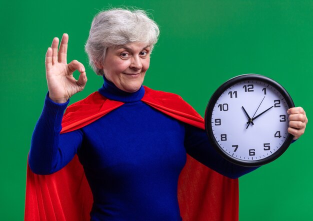 Mujer mayor superhéroe vistiendo capa roja sosteniendo el reloj de pared mirando a la cámara feliz y positivo mostrando signo ok de pie sobre fondo verde