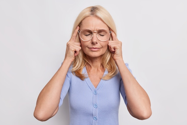 La mujer mayor tiene dolor de cabeza, toca las sienes con los ojos cerrados y la cara tensa sufre de migraña dolorosa, usa grandes lentes ópticos y un puente azul aislado sobre una pared blanca intenta enfocarse