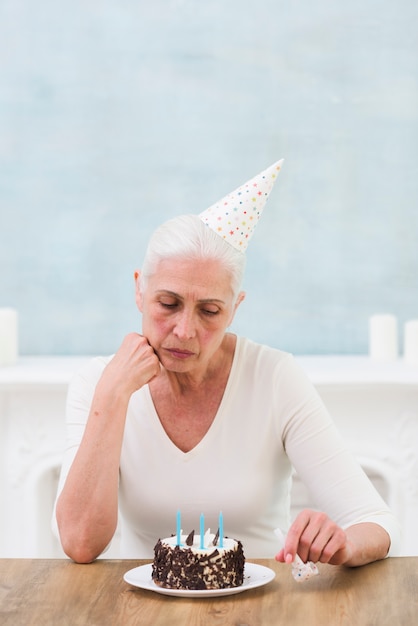 Mujer mayor triste que mira la torta de cumpleaños con la vela sobre la tabla