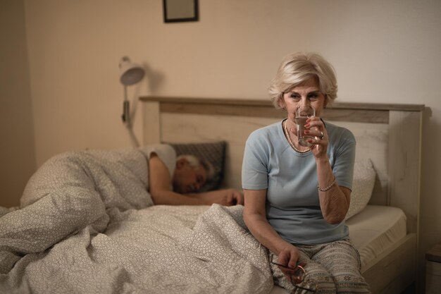 Mujer mayor con un vaso de agua en el dormitorio por la noche mientras su marido duerme