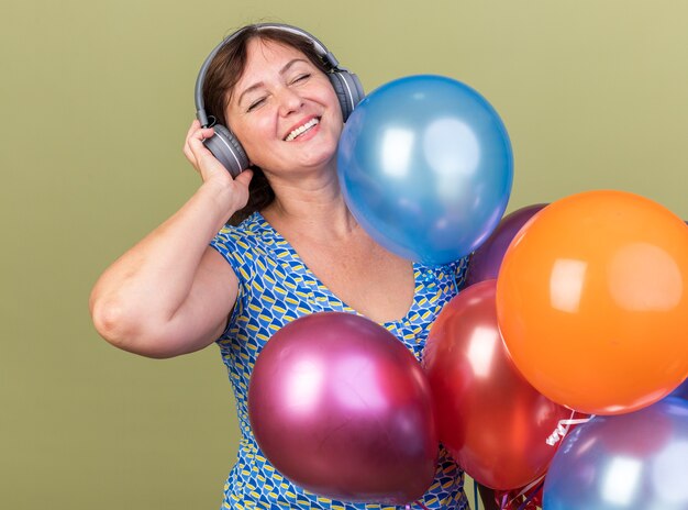 Mujer de mediana edad con auriculares y un montón de globos de colores feliz y alegre disfrutando de su música favorita