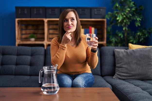 Mujer de mediana edad bebiendo un vaso de agua cara seria pensando en la pregunta con la mano en la barbilla pensativa sobre la idea confusa