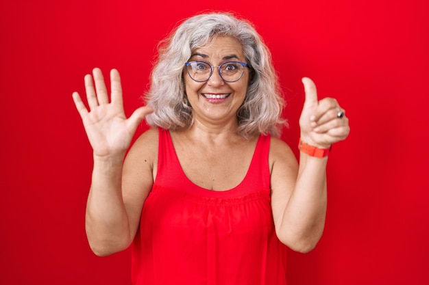 Mujer de mediana edad con cabello gris sobre fondo rojo que aparece y señala con los dedos número seis mientras sonríe confiada y feliz