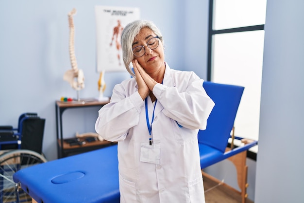 Mujer de mediana edad con cabello gris trabajando en la clínica de recuperación del dolor durmiendo cansada soñando y posando con las manos juntas mientras sonríe con los ojos cerrados.
