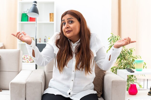 Mujer de mediana edad con camisa blanca y pantalón negro confundida extendiendo los brazos hacia los lados sin tener respuesta sentada en la silla en la sala de estar iluminada