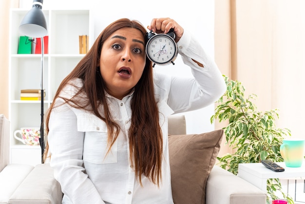 Mujer de mediana edad con camisa blanca y pantalón negro con reloj despertador mirando sorprendido y asombrado sentado en la silla en la sala de luz