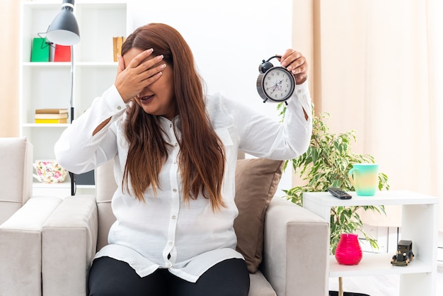 Mujer de mediana edad con camisa blanca y pantalón negro con reloj despertador que cubre los ojos con la mano witi sentada en la silla en la sala de luz