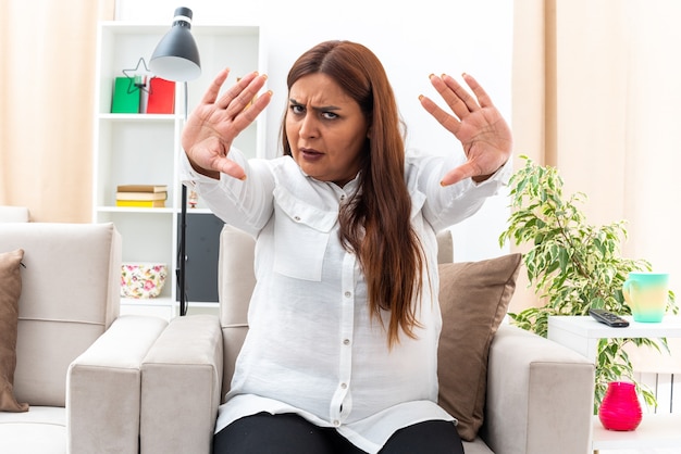 Mujer de mediana edad con camisa blanca y pantalón negro con rostro serio haciendo gesto de parada con las manos sentadas en la silla en la sala de luz