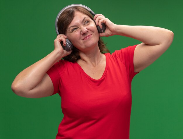 Mujer de mediana edad en camiseta roja con auriculares escuchando música disgustado de pie sobre la pared verde