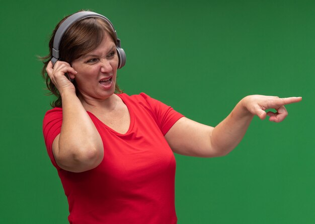 Mujer de mediana edad en camiseta roja con auriculares mirando a un lado disgustado apuntando con el dedo índice a algo parado sobre la pared verde