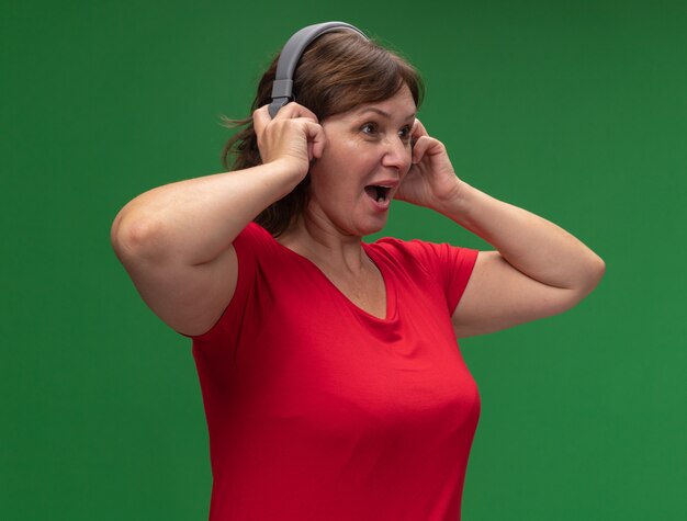 Mujer de mediana edad en camiseta roja con auriculares mirando a un lado feliz y sorprendido de pie sobre la pared verde