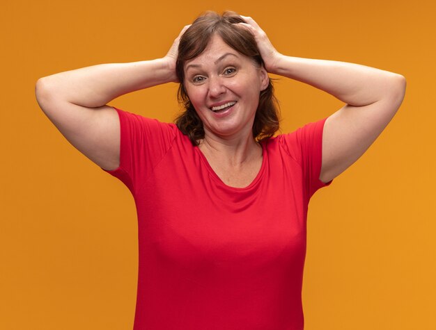 Mujer de mediana edad en camiseta roja feliz y positiva con las manos en la cabeza de pie sobre la pared naranja