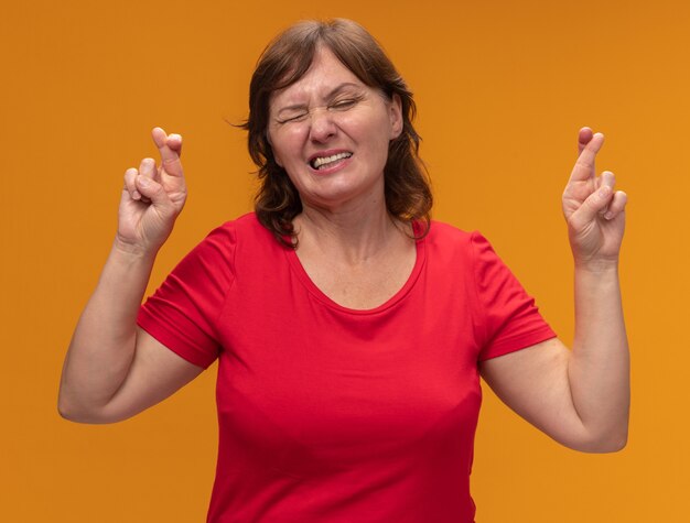 Mujer de mediana edad en camiseta roja haciendo un deseo deseable con expresión de esperanza cruzando los dedos con los ojos cerrados de pie sobre la pared naranja