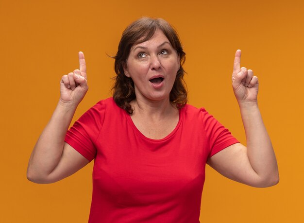 Mujer de mediana edad en camiseta roja mirando hacia arriba sonriendo señalando con el dedo índice de pie sobre la pared naranja