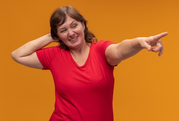 Mujer de mediana edad en camiseta roja mirando a un lado con la mano en la cabeza apuntando con el dedo índice a algo sonriente de pie sobre la pared naranja