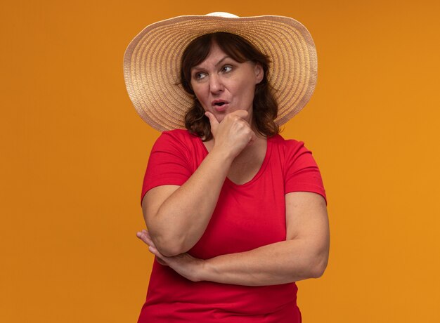 Mujer de mediana edad en camiseta roja y sombrero de verano mirando a un lado con expresión pensativa pensando de pie sobre la pared naranja