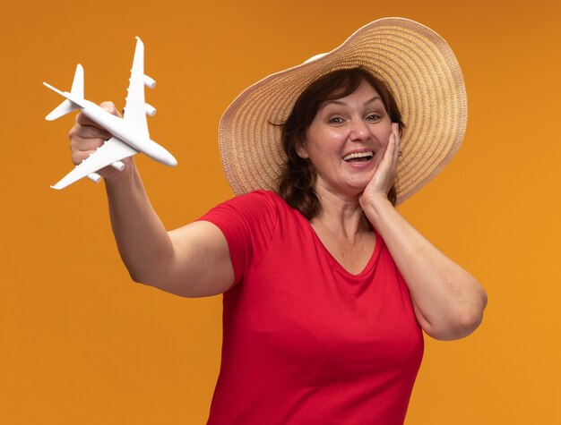Mujer de mediana edad en camiseta roja y sombrero de verano mostrando avión de juguete feliz y alegre sonriendo de pie sobre la pared naranja