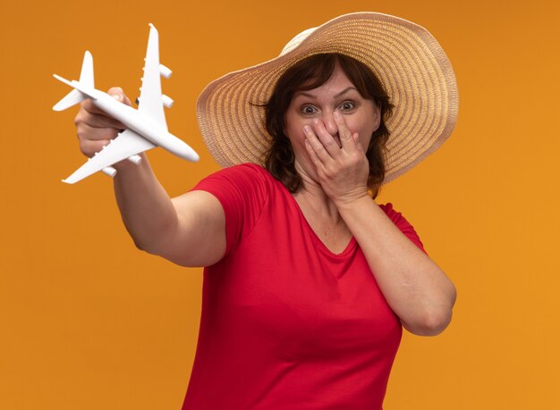 Mujer de mediana edad en camiseta roja y sombrero de verano mostrando un avión de juguete sorprendido cubriendo la boca con la mano de pie sobre la pared naranja