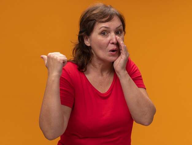 Mujer de mediana edad en camiseta roja susurrando un secreto con la mano cerca de la boca apuntando hacia atrás con el pulgar parado sobre la pared naranja