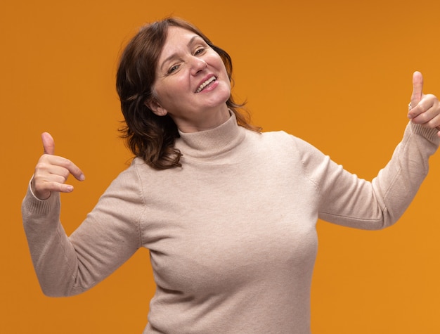 Foto gratuita mujer de mediana edad complacida en cuello alto beige apuntando a sí misma feliz y alegre de pie sobre la pared naranja
