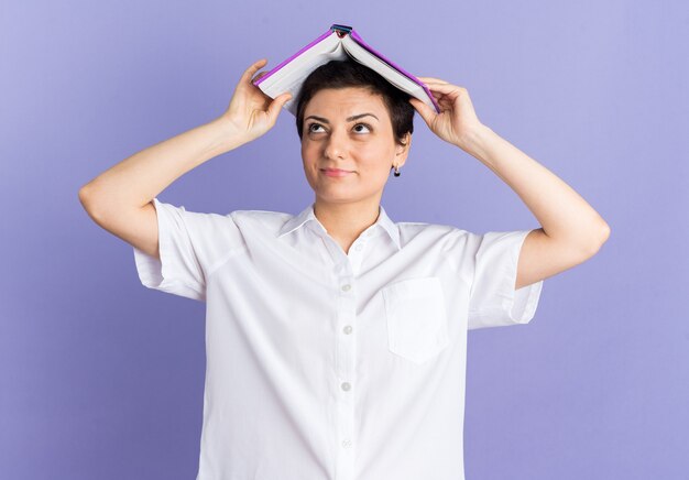 Mujer de mediana edad complacida sosteniendo el libro abierto en la cabeza mirando hacia arriba