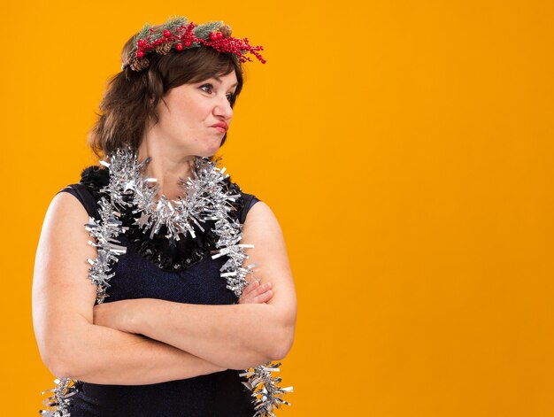 Mujer de mediana edad confundida con corona de Navidad y guirnalda de oropel alrededor del cuello de pie con postura cerrada mirando al lado aislado sobre fondo naranja con espacio de copia