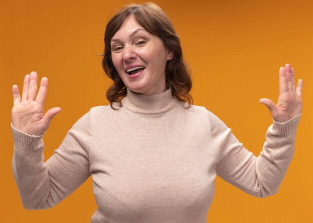 Mujer de mediana edad en cuello alto beige feliz y alegre sonriendo levantando las palmas en rendición de pie sobre la pared naranja