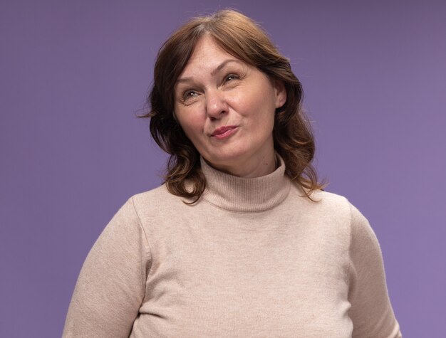 Mujer de mediana edad en cuello alto beige mirando hacia arriba disgustado de pie sobre la pared púrpura