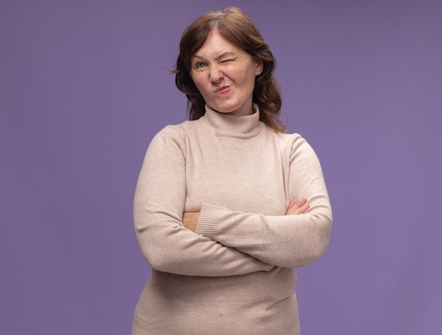 Mujer de mediana edad en cuello alto beige mirando a un lado con expresión escéptica con los brazos cruzados de pie sobre la pared púrpura