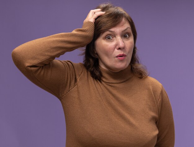 Foto gratuita mujer de mediana edad en cuello alto marrón confundida con la mano en la cabeza por error de pie sobre la pared púrpura