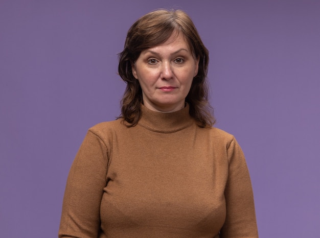 Foto gratuita mujer de mediana edad en cuello alto marrón con expresión de confianza de pie sobre la pared púrpura