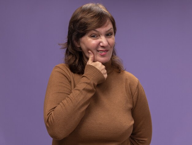 Mujer de mediana edad en cuello de tortuga marrón mirando confundido tocando su mejilla con dolor de muelas de pie sobre la pared púrpura