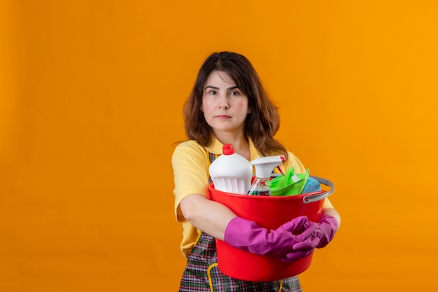 Mujer de mediana edad con delantal y guantes de goma sosteniendo un balde con herramientas de limpieza con expresión seria y segura en la cara de pie sobre la pared naranja