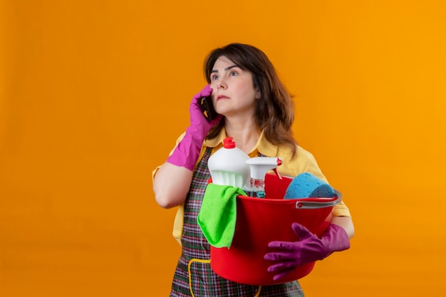 Mujer de mediana edad con delantal y guantes de goma sosteniendo un balde con herramientas de limpieza hablando por teléfono móvil con expresión seria en la cara de pie sobre la pared naranja