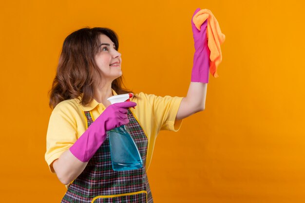 Mujer de mediana edad con delantal y guantes de goma con spray de limpieza y limpieza de alfombras con una sonrisa en la cara de pie sobre la pared naranja