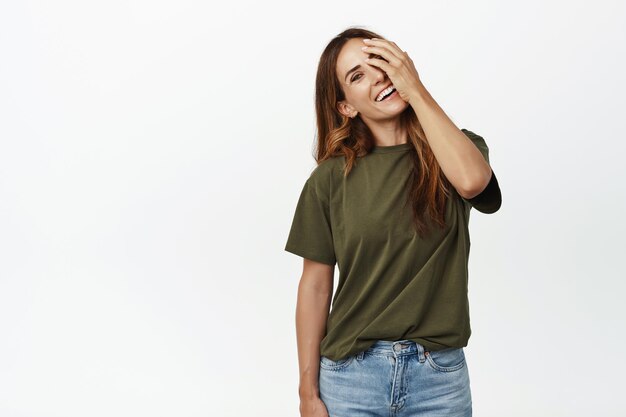 Mujer de mediana edad despreocupada con piel facial perfecta, sin arrugas, cabello largo saludable, tocándose la cara, cubriendo un ojo y sonriendo feliz a la cámara, fondo blanco