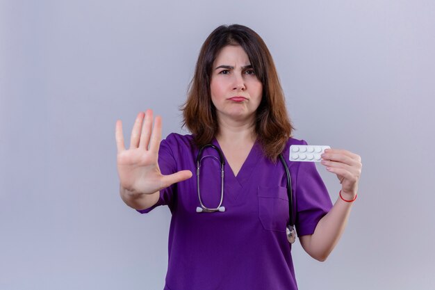 Mujer de mediana edad enfermera vistiendo uniforme médico y con estetoscopio sosteniendo blister con píldoras haciendo gesto de parada con la mano con el ceño fruncido sobre pared blanca