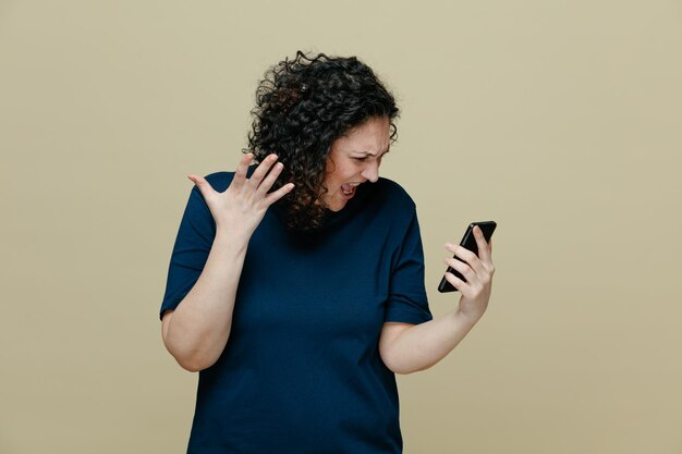 Mujer de mediana edad enojada con camiseta sosteniendo un teléfono móvil mirándolo mostrando gritos de mano vacíos aislados en un fondo verde oliva