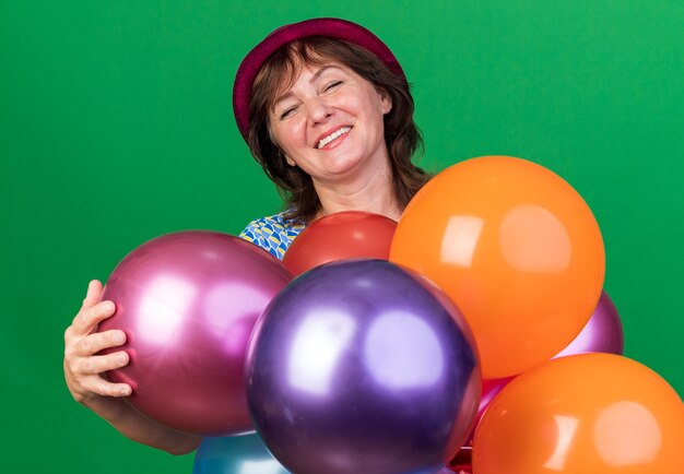 Mujer de mediana edad con gorro de fiesta con globos de colores feliz y feliz celebrando la fiesta de cumpleaños de pie sobre la pared verde