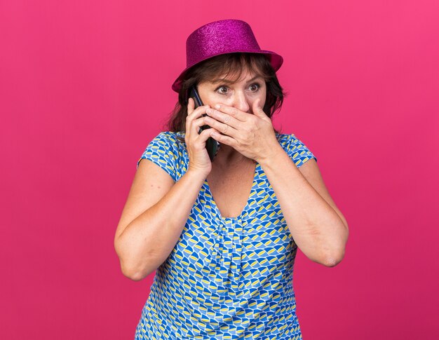 Mujer de mediana edad con gorro de fiesta mirando asombrado cubriendo la boca con la mano mientras habla por teléfono móvil